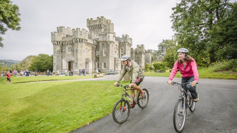 Two visitors biking downhill with Wray Castle and other visitors in the background